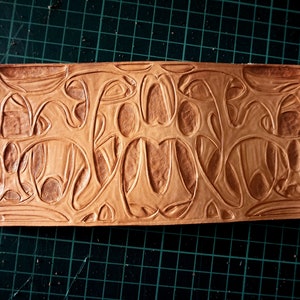 May include: A rectangular piece of light brown leather with an embossed organic design. The pattern features flowing, interconnected shapes and lines, creating a textured surface. The leather is set against a green cutting mat.