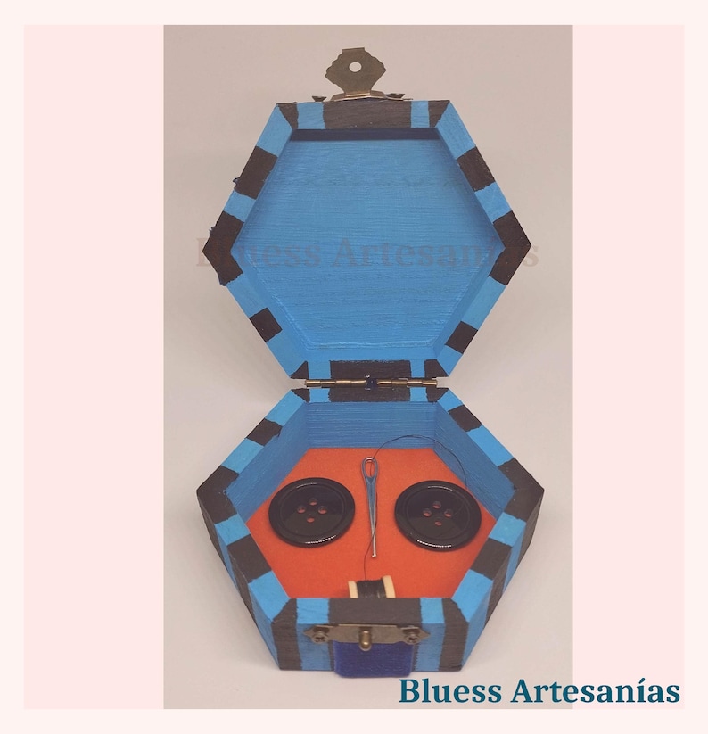Peut inclure: Bo&icirc;te &agrave; couture hexagonale en bois peinte en bleu avec des rayures noires. &Agrave; l'int&eacute;rieur, deux boutons noirs, une aiguille et du fil reposent sur une base orange. La bo&icirc;te est ouverte, r&eacute;v&eacute;lant son contenu. Le texte "Bluess Artesan&iacute;as" est visible.