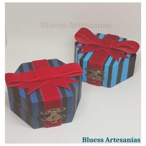 Op de afbeelding: Twee zeshoekige houten dozen met blauwe en zwarte strepen, afgewerkt met rode fluwelen strikken. De dozen hebben messing sluitingen en de tekst "Bluess Artesan&iacute;as" is zichtbaar.