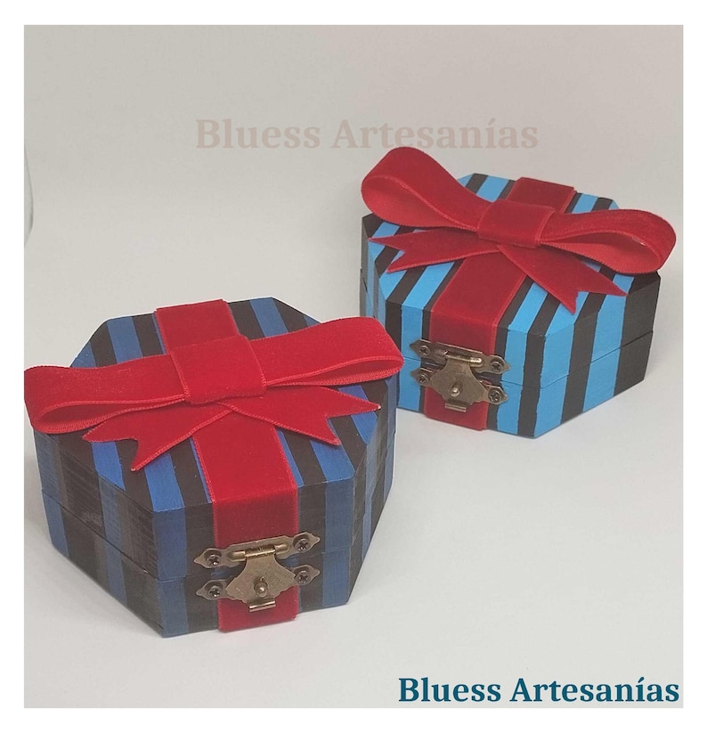 Op de afbeelding: Twee zeshoekige houten dozen met blauwe en zwarte strepen, afgewerkt met rode fluwelen strikken. De dozen hebben messing sluitingen en de tekst "Bluess Artesan&iacute;as" is zichtbaar.