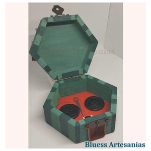 May include: Hexagonal wooden sewing box with a green and black striped exterior. The box is open, revealing a red interior with two black buttons and a needle and thread. The box has a small key and the text "Bluess Artesan&iacute;as".
