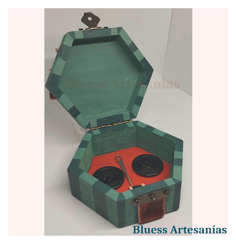 May include: Hexagonal wooden sewing box with a green and black striped exterior. The box is open, revealing a red interior with two black buttons and a needle and thread. The box has a small key and the text "Bluess Artesan&iacute;as".