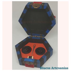 Op de afbeelding: Zeshoekige houten naaidoos met een zwart, blauw en rood kleurenschema. De doos bevat twee zwarte knopen, een naald en draad op een oranje viltbasis. De tekst "Bluess Artesan&iacute;as" is zichtbaar.