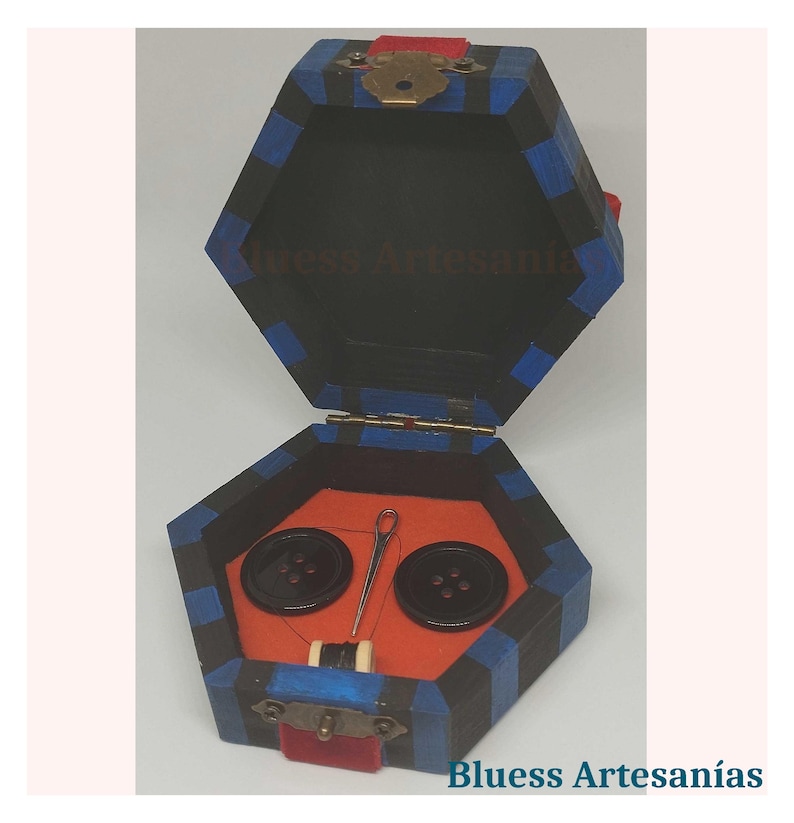 Op de afbeelding: Zeshoekige houten naaidoos met een zwart, blauw en rood kleurenschema. De doos bevat twee zwarte knopen, een naald en draad op een oranje viltbasis. De tekst "Bluess Artesan&iacute;as" is zichtbaar.