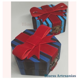 Op de afbeelding: Twee zeshoekige houten geschenkdozen met rode fluwelen strikken. De dozen zijn beschilderd met blauwe en zwarte strepen. De dozen hebben een kleine metalen sluiting. De tekst "Bluess Artesan&iacute;as" is zichtbaar.