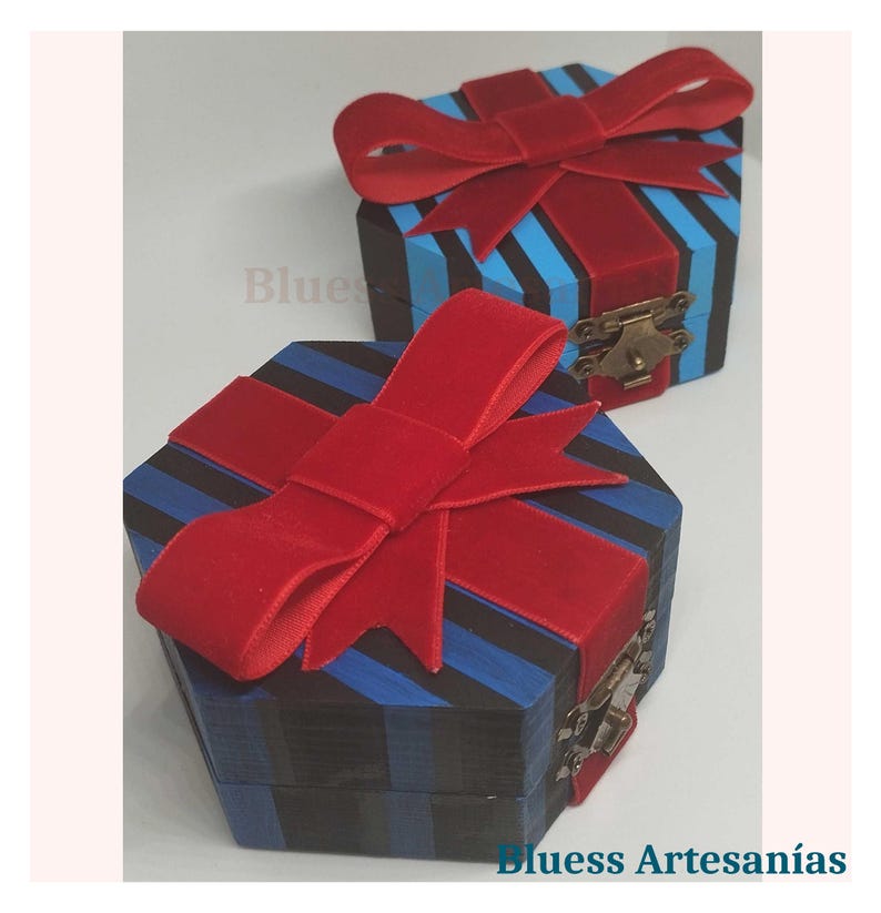 Op de afbeelding: Twee zeshoekige houten geschenkdozen met rode fluwelen strikken. De dozen zijn beschilderd met blauwe en zwarte strepen. De dozen hebben een kleine metalen sluiting. De tekst "Bluess Artesan&iacute;as" is zichtbaar.