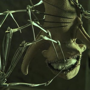 Pu&ograve; includere: Primo piano di una creatura inquietante, cucita, con una bocca piena di denti. La creatura &egrave; fatta di stoffa ed &egrave; attaccata a una struttura metallica.