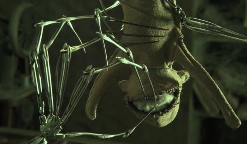 Pu&ograve; includere: Primo piano di una creatura inquietante, cucita, con una bocca piena di denti. La creatura &egrave; fatta di stoffa ed &egrave; attaccata a una struttura metallica.