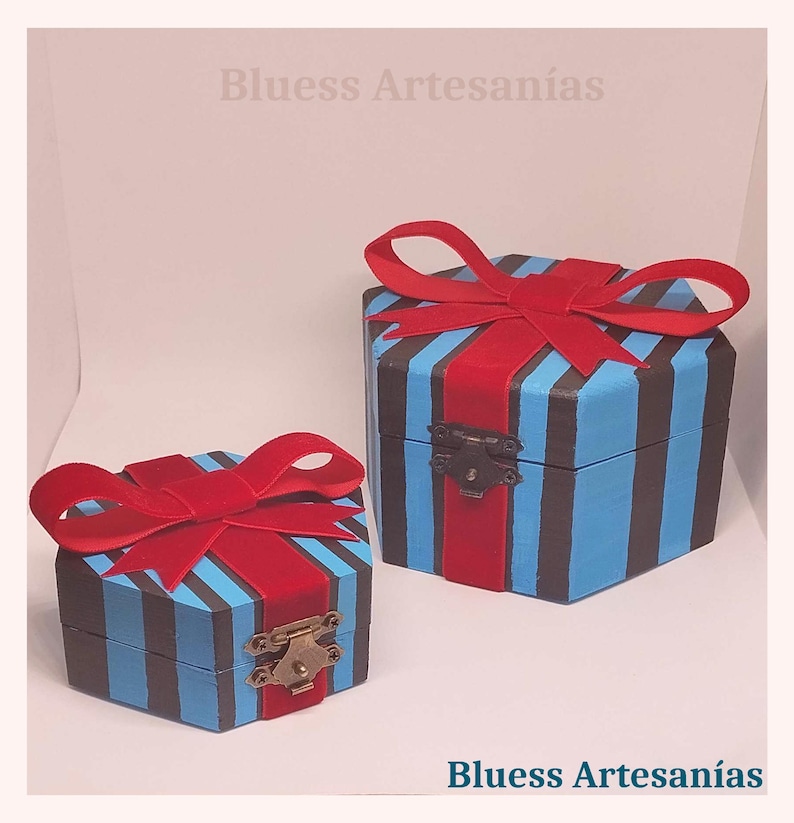 May include: Two hexagonal wooden boxes, painted with blue and black stripes, and adorned with red velvet bows. The boxes are secured with metal clasps. The text "Bluess Artesan&iacute;as" is visible.