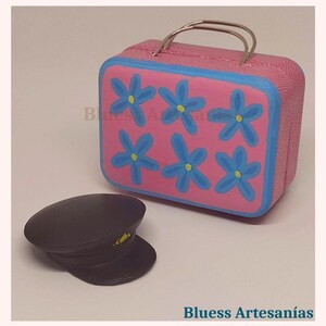 May include: A pink miniature suitcase with blue floral designs and a silver handle. Next to it is a black hat with a yellow accent. The text "Bluess Artesan&iacute;as" is visible.