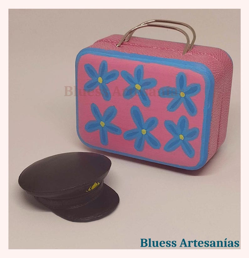May include: A pink miniature suitcase with blue floral designs and a silver handle. Next to it is a black hat with a yellow accent. The text "Bluess Artesan&iacute;as" is visible.