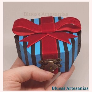 May include: An octagonal wooden box with blue and black stripes, topped with a red velvet bow. The box has a brass clasp. The text "Bluess Artesan&iacute;as" is visible.
