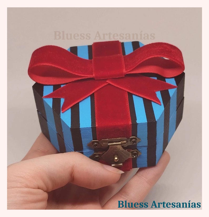 May include: An octagonal wooden box with blue and black stripes, topped with a red velvet bow. The box has a brass clasp. The text "Bluess Artesan&iacute;as" is visible.