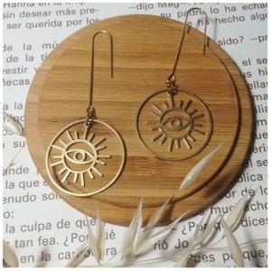 Puede incluir: Un par de pendientes de color dorado con un diseño circular que presenta un ojo estilizado con rayos de sol. Los pendientes están colgando de una superficie de madera.