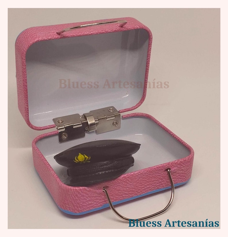 May include: A pink metal suitcase with a silver handle and hinges, open to reveal a black hat with a yellow emblem. The interior is white. The text "Bluess Artesan&iacute;as" is visible.