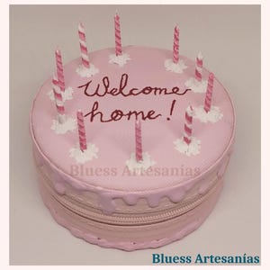 May include: A pink, round cake-shaped zippered bag with the words "Welcome home!" in burgundy script. The bag is decorated with pink and white striped candles and white frosting accents. The bottom of the bag has a zipper.