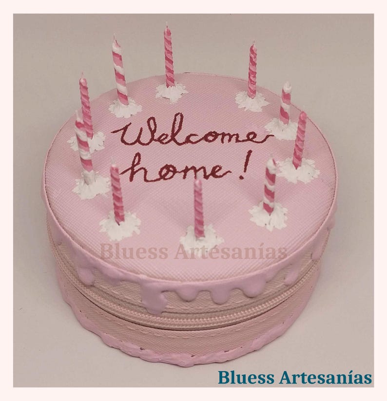 May include: A pink, round cake-shaped zippered bag with the words "Welcome home!" in burgundy script. The bag is decorated with pink and white striped candles and white frosting accents. The bottom of the bag has a zipper.