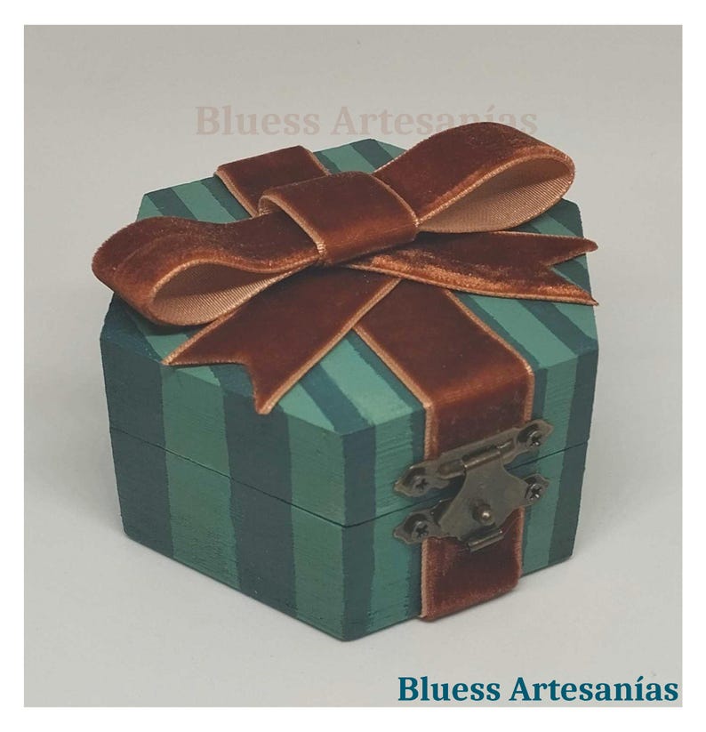 May include: A small, hexagonal wooden box painted teal with vertical stripes and a brown velvet ribbon tied in a bow. The box has a metal clasp and the text "Bluess Artesan&iacute;as".