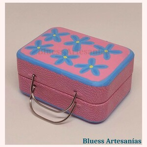 May include: A pink, rectangular box with a blue rim and a silver handle. The top of the box is decorated with blue flower designs. The text "Bluess Artesan&iacute;as" is visible on the bottom of the image.