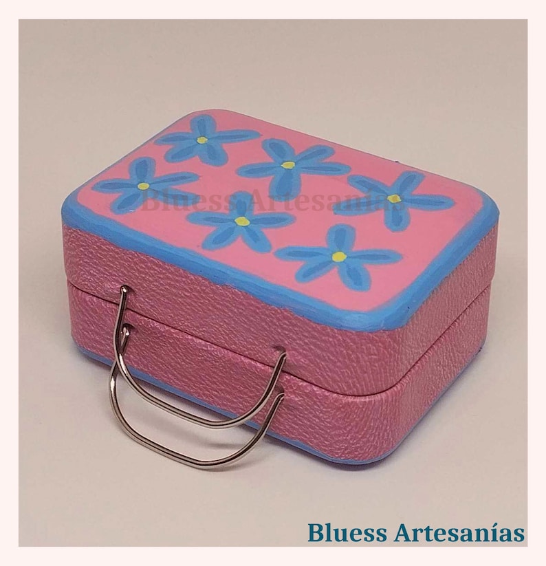 May include: A pink, rectangular box with a blue rim and a silver handle. The top of the box is decorated with blue flower designs. The text "Bluess Artesan&iacute;as" is visible on the bottom of the image.