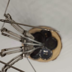 Pu&ograve; includere: Un set di aghi da cucito argentati disposti in un motivo circolare su una base di legno. Gli aghi sono tutti puntati verso l'alto e sono tenuti in posizione da un anello di metallo. La base &egrave; in legno marrone chiaro e ha una finitura naturale.