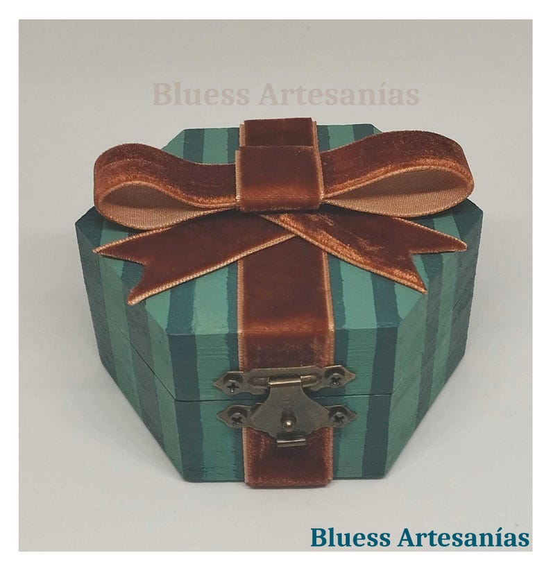 May include: A teal and green striped wooden box with a brown velvet ribbon and bow. The box has a metal clasp and the text "Bluess Artesan&iacute;as". This decorative box is suitable for small gifts or keepsakes.