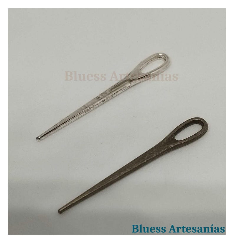 Op de afbeelding: Twee metalen splitpennen, &eacute;&eacute;n zilverkleurig en &eacute;&eacute;n bronskleurig, worden getoond tegen een lichte achtergrond. De pennen hebben een lus aan &eacute;&eacute;n uiteinde en een punt aan het andere. De tekst "Bluess Artesan&iacute;as" is zichtbaar.