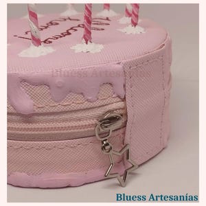 May include: A pink, round cake-shaped cosmetic bag with a zip closure and a star-shaped charm. The bag has pink frosting details and decorative candles. The text "Blusess Artesan&iacute;as" is visible.