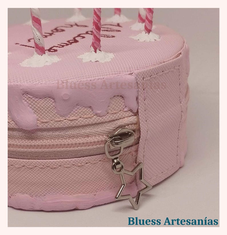 May include: A pink, round cake-shaped cosmetic bag with a zip closure and a star-shaped charm. The bag has pink frosting details and decorative candles. The text "Blusess Artesan&iacute;as" is visible.