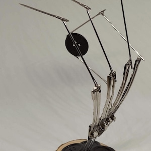 Pu&ograve; includere: Una scultura in metallo di una mano realizzata con spille da balia e bottoni. La mano tiene un mucchio di bottoni neri su una base di legno. La scultura &egrave; intitolata "BluessArtesanias".