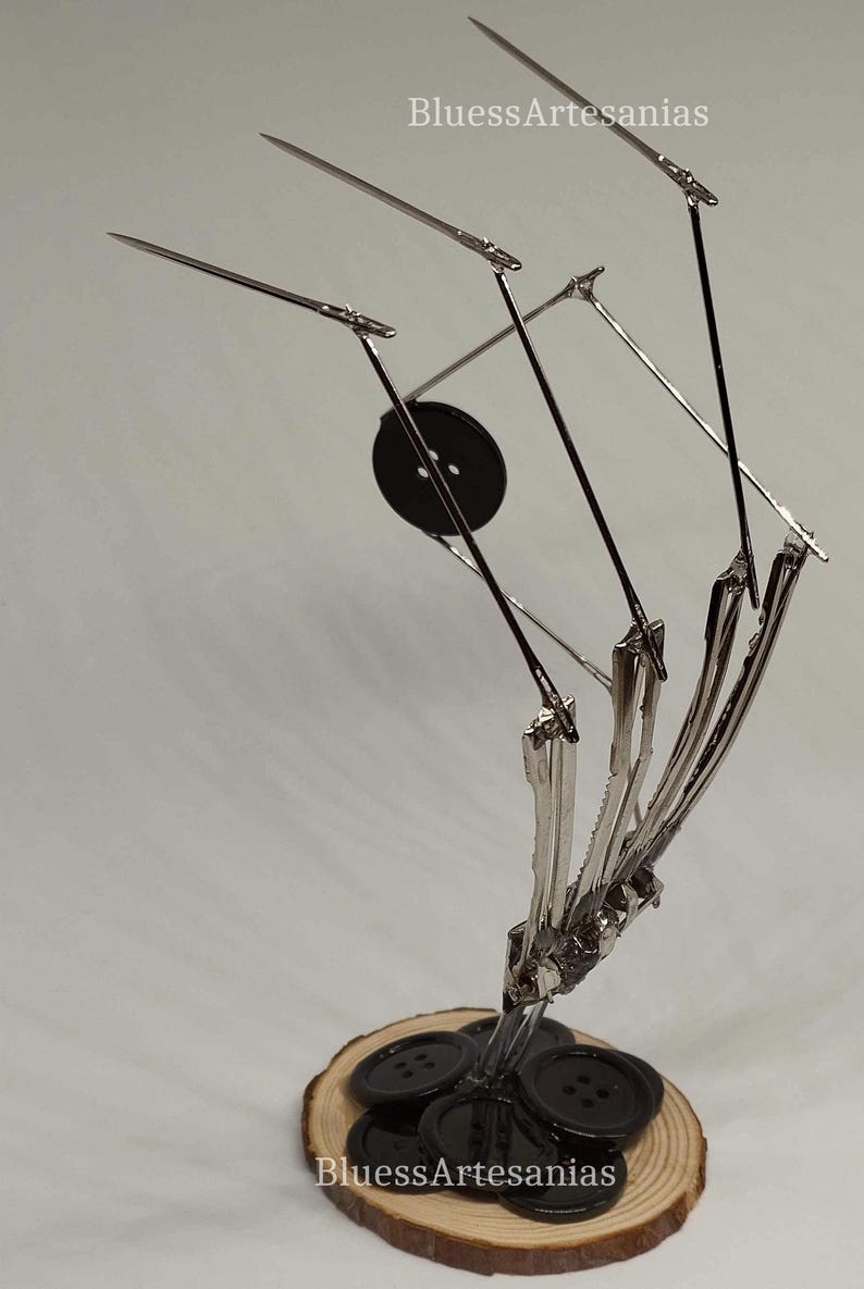 Pu&ograve; includere: Una scultura in metallo di una mano realizzata con spille da balia e bottoni. La mano tiene un mucchio di bottoni neri su una base di legno. La scultura &egrave; intitolata "BluessArtesanias".