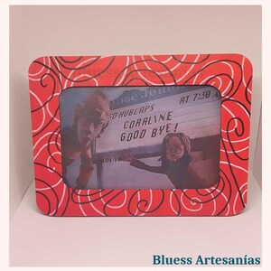 May include: A rectangular red picture frame with a white swirl design. The frame holds a black and white image of two people and a sign that reads "CORALINE GOOD BYE!". The frame is on a white surface.