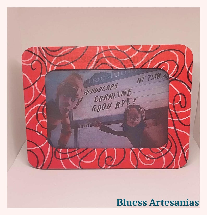 May include: A rectangular red picture frame with a white swirl design. The frame holds a black and white image of two people and a sign that reads "CORALINE GOOD BYE!". The frame is on a white surface.
