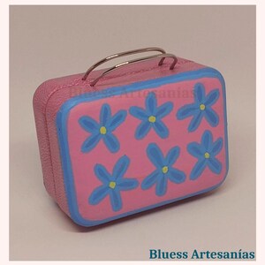 May include: A pink, rectangular miniature suitcase with a blue border and six blue flower designs with yellow centers. The suitcase has silver handles and the text "Bluess Artesan&iacute;as".
