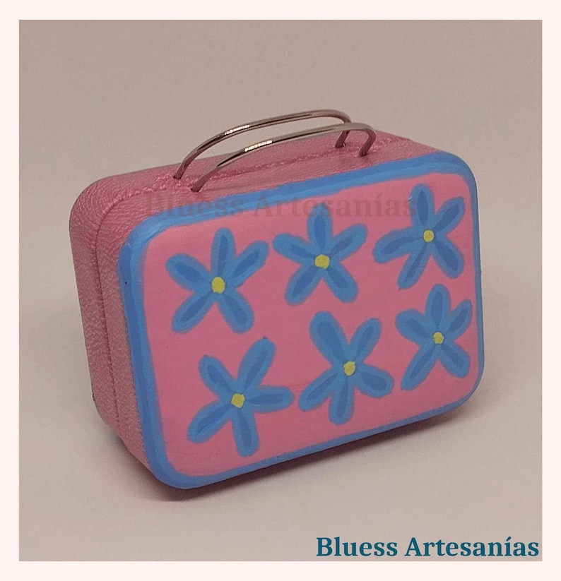 May include: A pink, rectangular miniature suitcase with a blue border and six blue flower designs with yellow centers. The suitcase has silver handles and the text "Bluess Artesan&iacute;as".