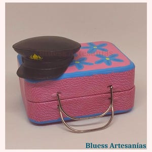 May include: A miniature pink suitcase with blue trim and blue flower accents, paired with a black hat. The suitcase has a silver handle. The text "Bluess Artesan&iacute;as" is visible at the bottom of the image.