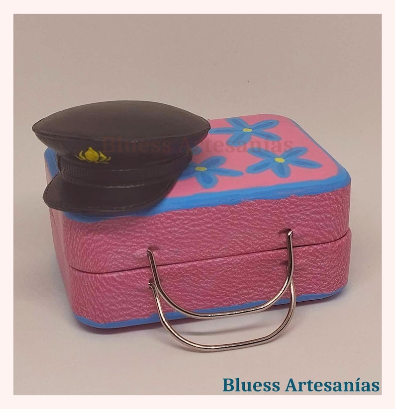 May include: A miniature pink suitcase with blue trim and blue flower accents, paired with a black hat. The suitcase has a silver handle. The text "Bluess Artesan&iacute;as" is visible at the bottom of the image.