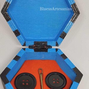 May include: Hexagonal wooden sewing box, painted blue with black stripes, open to reveal an orange interior. Inside are two large black buttons, a needle, and a spool of thread. The text "BluessArtesanias" is visible.