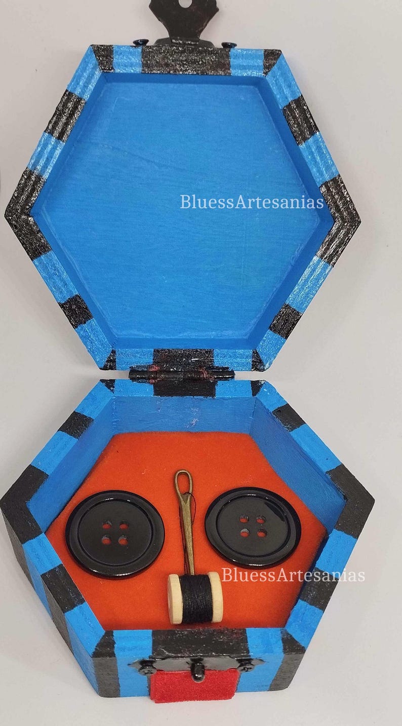 May include: Hexagonal wooden sewing box, painted blue with black stripes, open to reveal an orange interior. Inside are two large black buttons, a needle, and a spool of thread. The text "BluessArtesanias" is visible.
