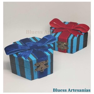 Peut inclure: Deux petites bo&icirc;tes d&eacute;coratives en bois avec couvercles &agrave; charni&egrave;res. Les bo&icirc;tes sont peintes de rayures verticales bleues et noires et surmont&eacute;es de n&oelig;uds en velours. Une bo&icirc;te a un n&oelig;ud bleu, l'autre un n&oelig;ud rouge. Les bo&icirc;tes ont des fermoirs en m&eacute;tal. Le texte "Bluess Artesan&iacute;as" est visible.
