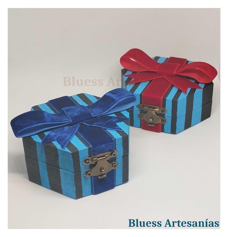 Peut inclure: Deux petites bo&icirc;tes d&eacute;coratives en bois avec couvercles &agrave; charni&egrave;res. Les bo&icirc;tes sont peintes de rayures verticales bleues et noires et surmont&eacute;es de n&oelig;uds en velours. Une bo&icirc;te a un n&oelig;ud bleu, l'autre un n&oelig;ud rouge. Les bo&icirc;tes ont des fermoirs en m&eacute;tal. Le texte "Bluess Artesan&iacute;as" est visible.