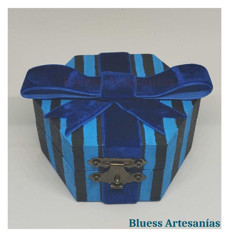 Peut inclure: Une bo&icirc;te en bois ray&eacute;e bleue et noire avec un n&oelig;ud en velours. La bo&icirc;te a un fermoir en m&eacute;tal et le texte "Bluess Artesan&iacute;as" en bas. Le n&oelig;ud est bleu fonc&eacute;.