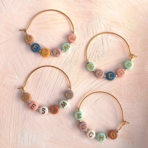 Op de afbeelding: Vier gouden oorringen met kleurrijke kralen die "BESAME MUCHO" spellen. De kralen zijn in pasteltinten van blauw, roze, geel en bruin. De oorbellen worden gepresenteerd op een roze, getextureerd oppervlak.