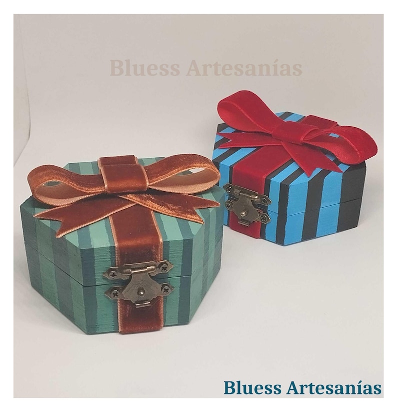 May include: Two decorative wooden boxes with lids. One is teal with brown stripes and a brown velvet bow. The other is blue with red stripes and a red velvet bow. Both have metal clasps. The text "Bluess Artesan&iacute;as" is visible.