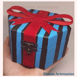 May include: A blue and black striped wooden box with a red ribbon tied on top. The box is held in a hand. The box has a metal clasp. The text "Bluess Artesan&iacute;as" is visible.