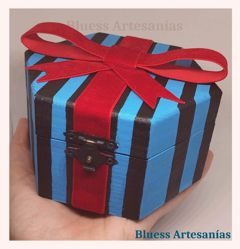 May include: A blue and black striped wooden box with a red ribbon tied on top. The box is held in a hand. The box has a metal clasp. The text "Bluess Artesan&iacute;as" is visible.