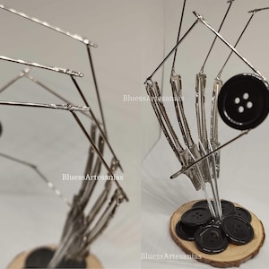 Pu&ograve; includere: Un bottone nero con quattro fori &egrave; tenuto in posizione da una scultura metallica realizzata con mollette per capelli. La scultura &egrave; montata su una base di legno.
