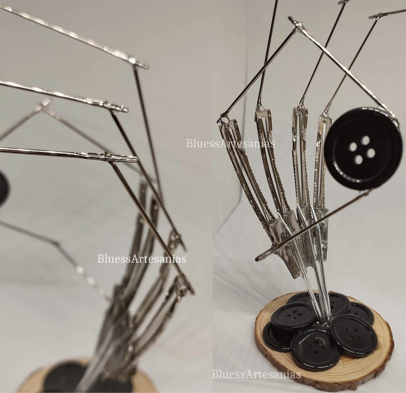 Pu&ograve; includere: Un bottone nero con quattro fori &egrave; tenuto in posizione da una scultura metallica realizzata con mollette per capelli. La scultura &egrave; montata su una base di legno.