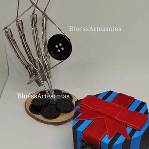 May include: A decorative display featuring a metal button holder with black buttons on a wooden base, and a blue and black striped gift box with a red bow. The text "BluessArtesanias" is visible.