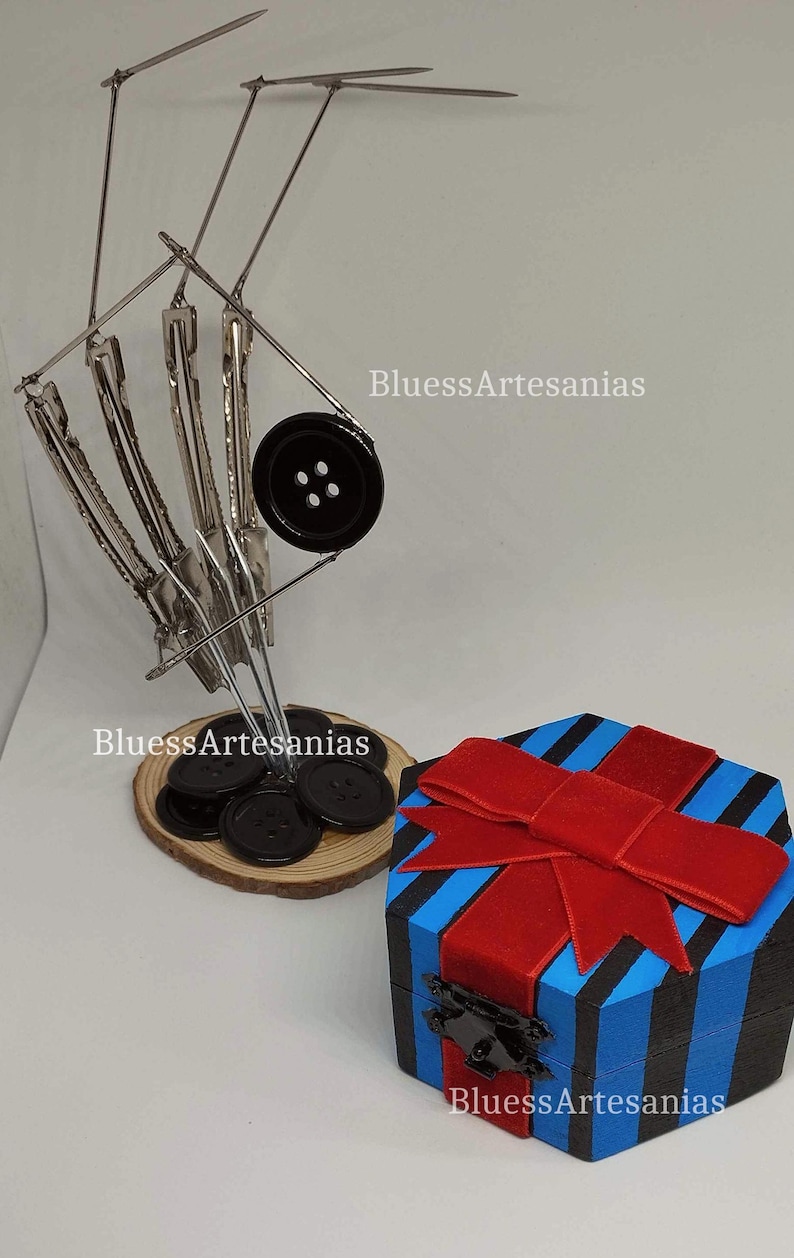 May include: A decorative display featuring a metal button holder with black buttons on a wooden base, and a blue and black striped gift box with a red bow. The text "BluessArtesanias" is visible.